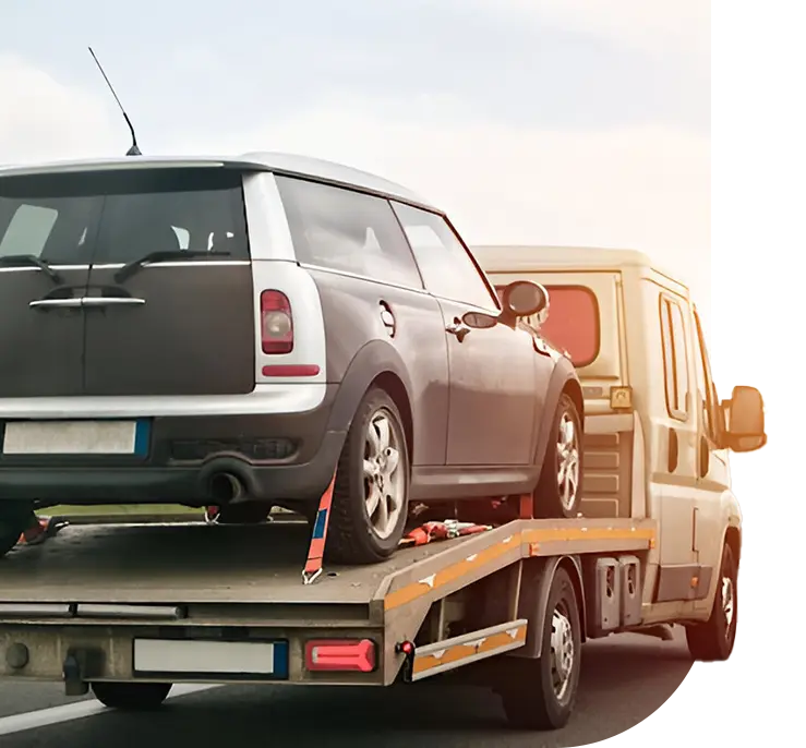 A grey car loaded on a tow truck's flatbed, with a light van in the foreground, set against a soft sunset background.