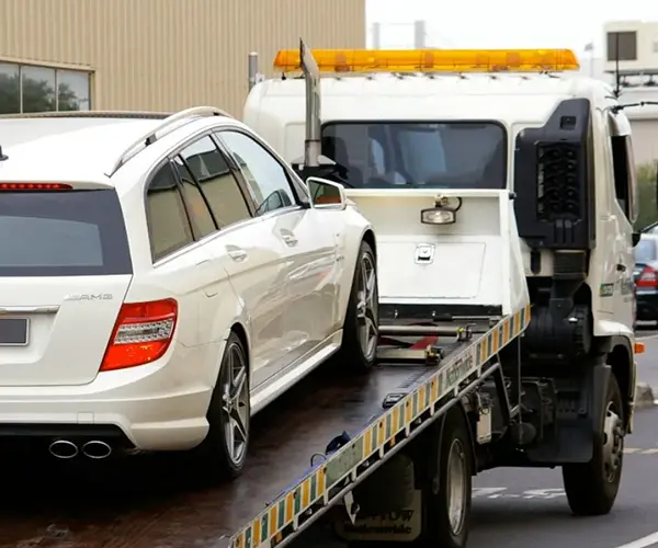 A white Mercedes-Benz station wagon being loaded onto a tow truck, with an industrial background and emergency lights flashing.