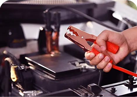 A hand holds a red jumper cable clamp above a car battery, preparing to jump-start the engine.