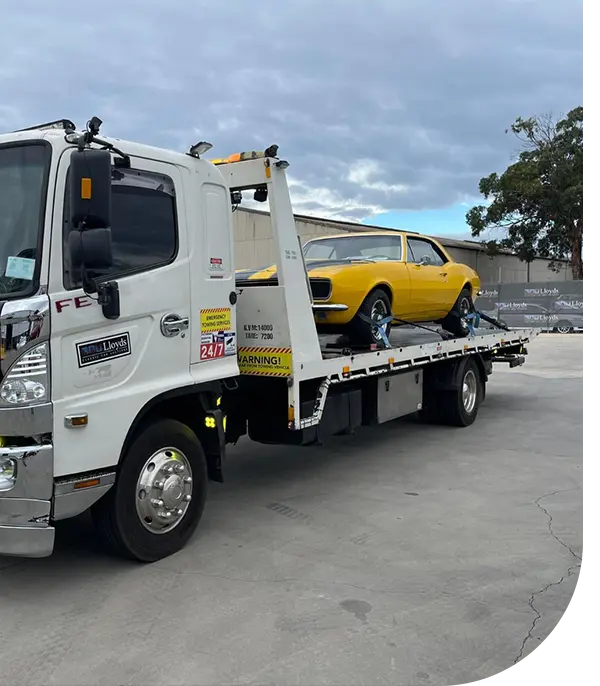 A white tow truck transports a classic yellow car, parked securely on the flatbed, under a cloudy sky in an industrial area.
