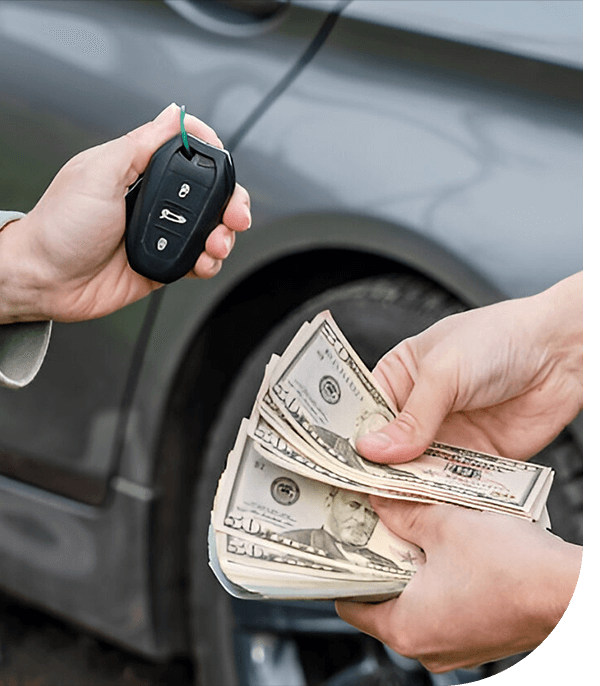 A person hands over car keys while receiving a stack of cash in front of a grey vehicle.