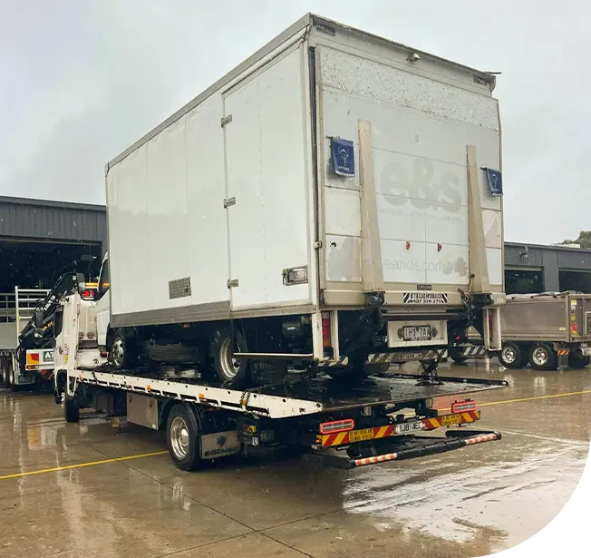 A tow truck lifts a white delivery van during rain, parked in an industrial area with several trailers in the background.