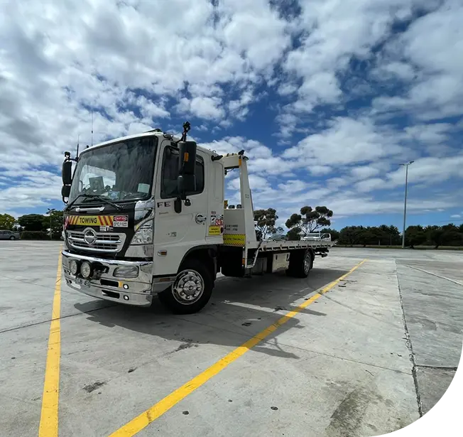 A white tow truck parked in a lot under a partly cloudy sky, showcasing its flatbed and towing equipment.