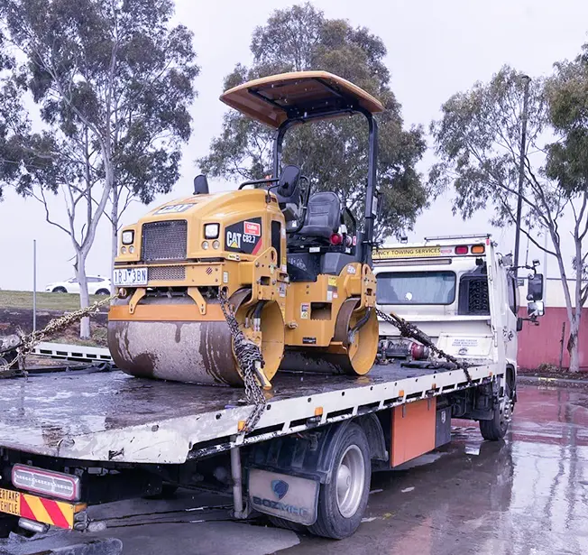 A yellow CAT roller is secured on a tow truck's flatbed, surrounded by trees in a rainy landscape.
