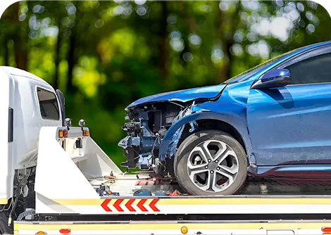 A damaged blue car with a crumpled front end being loaded onto a tow truck against a blurred green background.