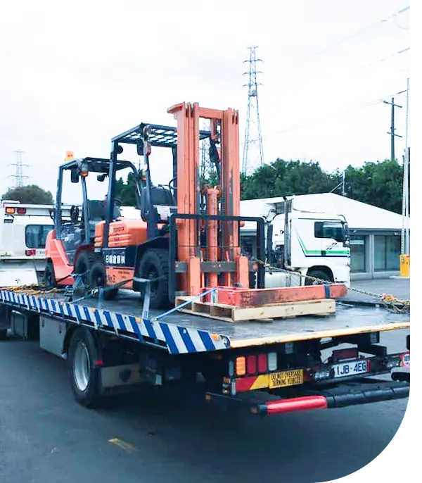 A flatbed truck transports an orange forklift, with power lines and other vehicles visible in the background.