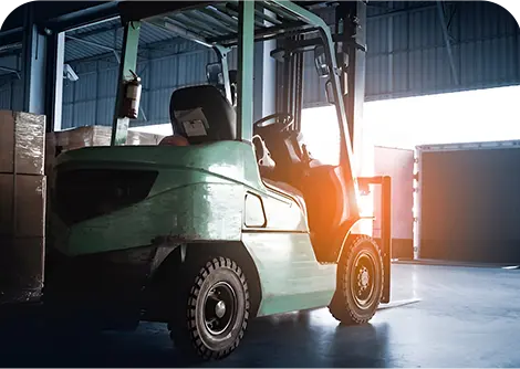 A green forklift is parked in a warehouse, illuminated by sunlight streaming through an open door, casting shadows on the floor.