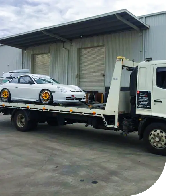 A white sports car with yellow rims is loaded onto a tow truck outside a storage warehouse under a cloudy sky.
