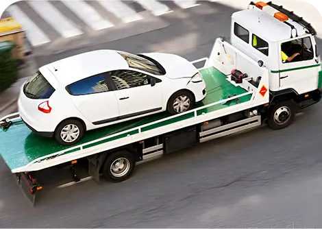 A white car is being loaded onto a flatbed tow truck, which is driving down a city street.