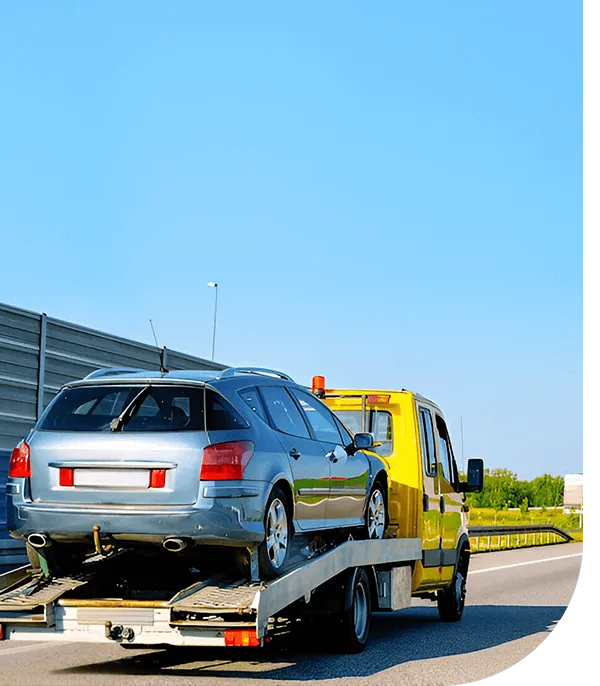 A yellow tow truck transporting a silver car on a flatbed along a sunny highway with clear blue skies.