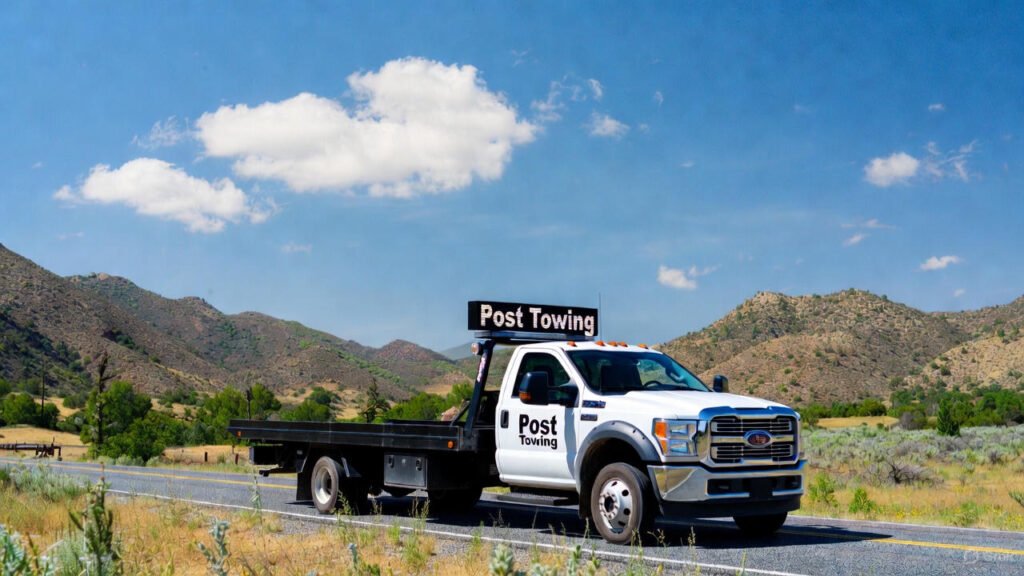 What to Do After Your Car Is Towed: Your Complete Post-Towing Checklist 2 A white flatbed tow truck with "Post Towing" signage drives along a scenic highway, surrounded by mountains and greenery under a blue sky. Post-Towing Checklist