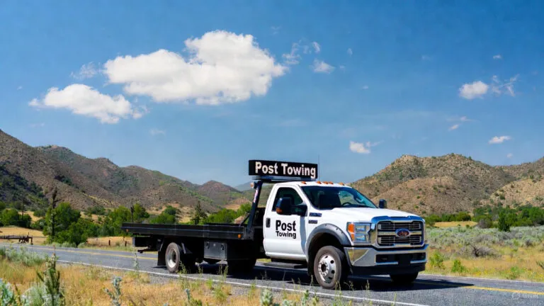 A white flatbed tow truck with "Post Towing" signage drives along a scenic highway, surrounded by mountains and greenery under a blue sky. Post-Towing Checklist